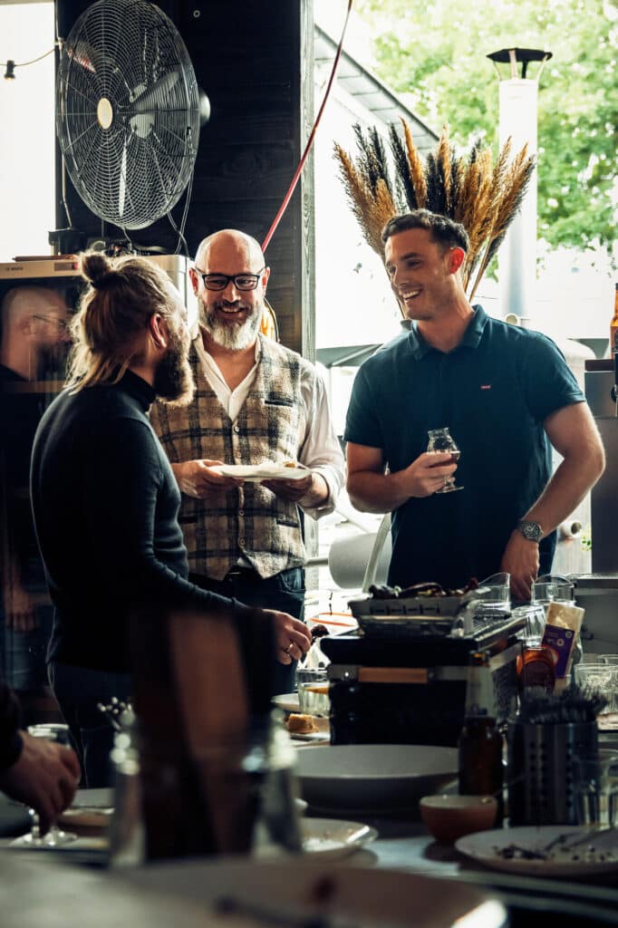 Drie mannen praten en lachen rondom een tafel met eten.